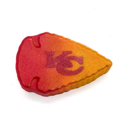 A bath bomb shaped like an arrowhead with a gradient color scheme transitioning from red to yellow. The initials "KC" are prominently displayed in a darker shade, symbolizing the Kansas City team