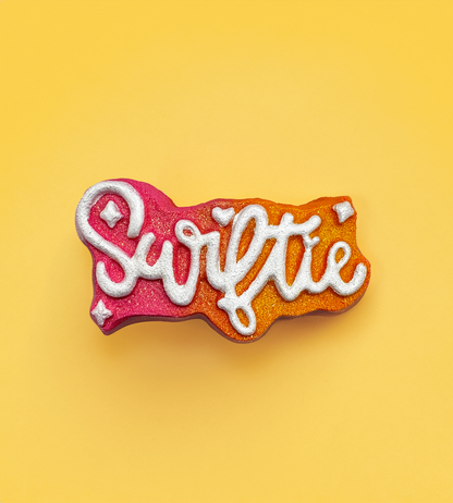 A bath bomb shaped like a speech bubble with the word "Swiftie" written in cursive glittery white font. The bath bomb features a gradient color scheme transitioning from bright pink on the left to warm orange on the right. The background is a solid pink color.